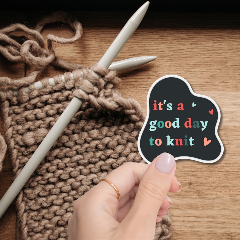 May include: A black and white sticker with the text "it's a good day to knit" in colorful letters. The sticker has a heart shape at the top and bottom. A hand is holding the sticker in front of a brown knitted fabric.