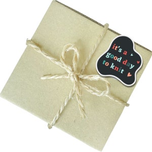 May include: A brown paper package tied with twine and a black sticker that says "it's a good day to knit" with red, pink, and teal lettering and small hearts.