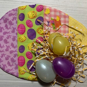 May include: A purple, green, pink, and yellow fabric Easter egg basket with a bunny pattern, a checkered pattern, and an egg pattern. The basket is filled with yellow, purple, and green plastic Easter eggs and yellow shredded paper.