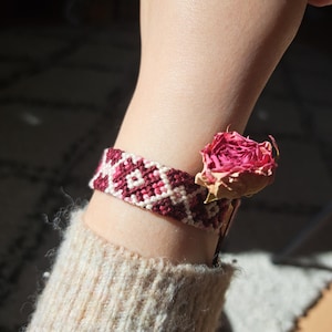 Könnte beinhalten: Handgefertigtes Freundschaftsarmband in Burgunderrot und Creme mit geometrischem Muster. Eine getrocknete rosa Rose liegt neben dem Armband. Das Armband wird am Handgelenk getragen, ein beiger Ärmel ist sichtbar.