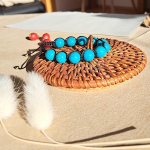 Könnte beinhalten: Zwei türkisfarbene Armbänder mit schwarzen Schnüren werden auf einem kleinen, runden, gewebten Tablett aus Korbgeflecht präsentiert. Die Armbänder haben große, runde, leuchtend blaue Perlen. Der Hintergrund umfasst weiße, flauschige Dekorationselemente und kleine rote Beeren.