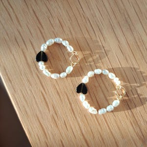 Könnte beinhalten: Zwei Perlenarmbänder mit einem schwarzen herzförmigen Anhänger und einem goldfarbenen Verschluss. Die Armbänder liegen auf einer hellbraunen Holzoberfläche. Die Perlen sind weiß und oval geformt.