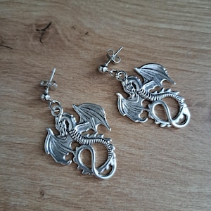 Könnte beinhalten: Ein Paar silberfarbene Drachenohrringe. Jeder Ohrring hat ein detailliertes Drachendesign mit ausgebreiteten Flügeln und einem eingerollten Schwanz. Die Ohrringe haben einen Steckerverschluss und werden auf einer Holzoberfläche präsentiert.