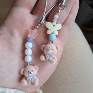 Könnte beinhalten: Zwei Perlen-Telefonanhänger. Einer hat einen rosa Stern, lila und weiße Perlen und einen kleinen Teddybären mit einer blauen Schleife. Der andere hat einen Schmetterling, eine blaue Perle und einen Teddybären. Beide haben silberfarbene Hardware.