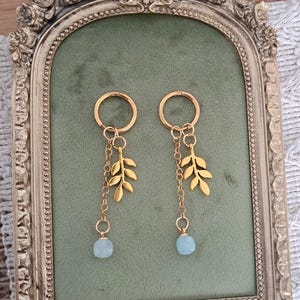 Könnte beinhalten: Ein Paar goldfarbene Ohrringe mit Blattdesign und einem hellblauen Edelstein. Die Ohrringe hängen an einem goldenen Ring und werden auf einem grünen Samthintergrund in einem dekorativen Rahmen präsentiert.