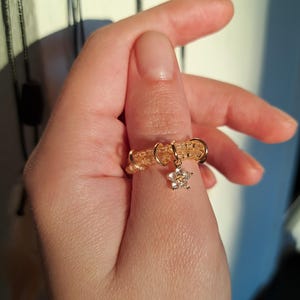 Könnte beinhalten: Ein goldfarbener Perlenring mit einem kleinen, baumelnden Blumenanhänger. Der Ring wird an einem Finger getragen und zeigt die komplizierte Perlenarbeit und den zarten Blumenanhänger. Das Design des Rings ist schlicht, aber elegant.