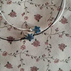 Könnte beinhalten: Zwei zarte Halsketten mit sternförmigen blauen Perlen. Eine Halskette hat eine weiße Kordel, die andere eine schwarze Kordel. Die Perlen haben eine durchscheinende, irisierende Qualität. Der Hintergrund ist ein Blumenmuster in Rosa und Grün.