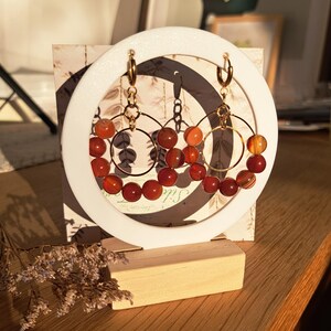 Könnte beinhalten: Goldfarbene Creolen mit rot-orangen Perlen, präsentiert auf einem weißen, kreisförmigen Ständer mit Holzsockel. Der Hintergrund zeigt eine gemusterte Karte und eine Holzoberfläche.