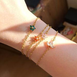 Könnte beinhalten: Vier goldfarbene Kettenarmbänder, jedes mit einem anderen Blumen- oder Schmetterlingsanhänger. Die Anhänger sind mit grünen, orangefarbenen, rosa und klaren Edelsteinen besetzt. Die Armbänder werden am Handgelenk einer Person präsentiert.