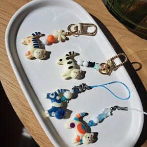 Pode incluir: Quatro pingentes em forma de gato com padrões coloridos, incluindo manchas ou listras laranjas e azuis. Cada pingente está preso a um fio de contas com formas de estrelas e esferas, terminando em um fecho de metal dourado ou um cordão fino.