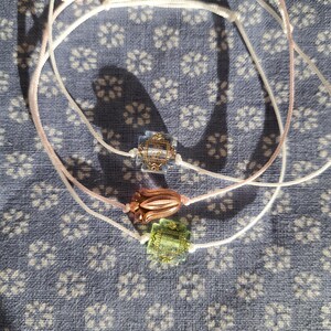 Könnte beinhalten: Drei zarte Armbänder mit dekorativen Perlen. Ein Armband hat eine blaue Perle, eines eine kupferfarbene Perle und eines eine grüne Perle. Die Armbänder befinden sich auf einem blauen Stoff mit Blumenmuster.