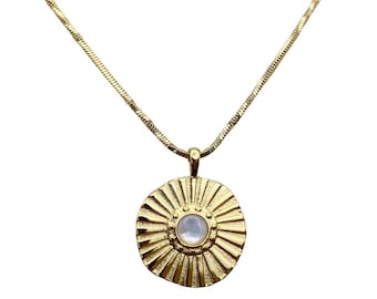 May include: A gold-colored necklace with a circular pendant. The pendant features a sunburst design with a central, iridescent, round stone. The chain is a delicate, textured gold. The necklace is displayed against a white background.