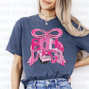 May include: A navy blue t-shirt featuring a pink bow and the word "BULLDOGS" in pink letters. The bulldog graphic is wearing pink star-shaped sunglasses. The shirt is a comfortable, casual style.