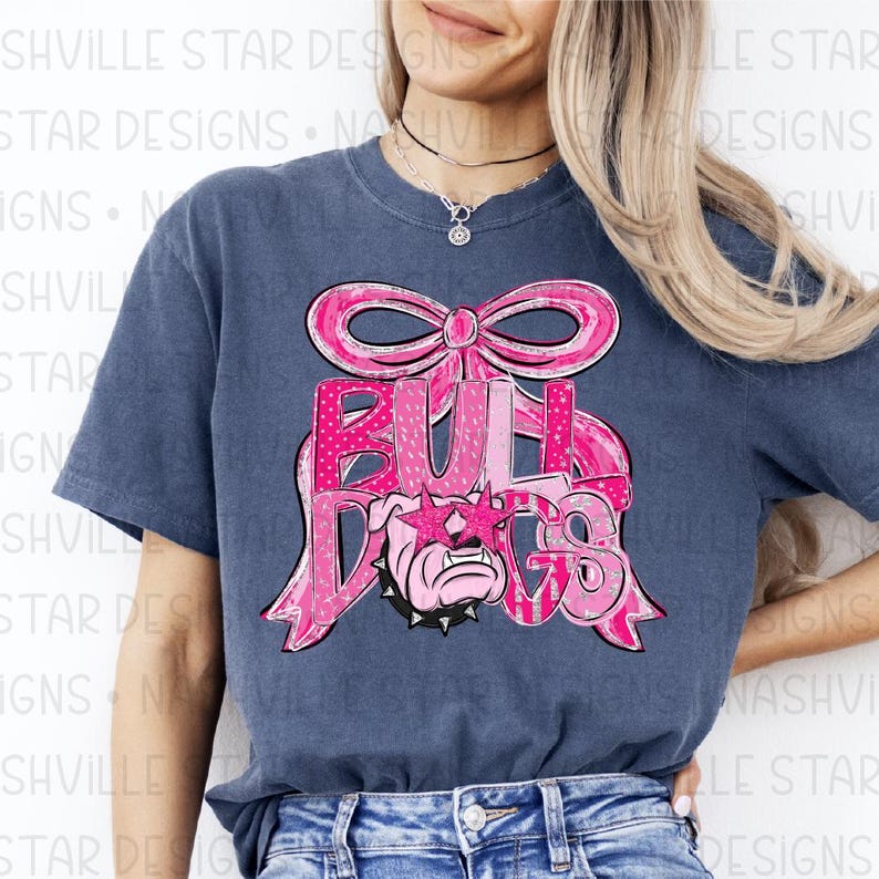 May include: A navy blue t-shirt featuring a pink bow and the word "BULLDOGS" in pink letters. The bulldog graphic is wearing pink star-shaped sunglasses. The shirt is a comfortable, casual style.