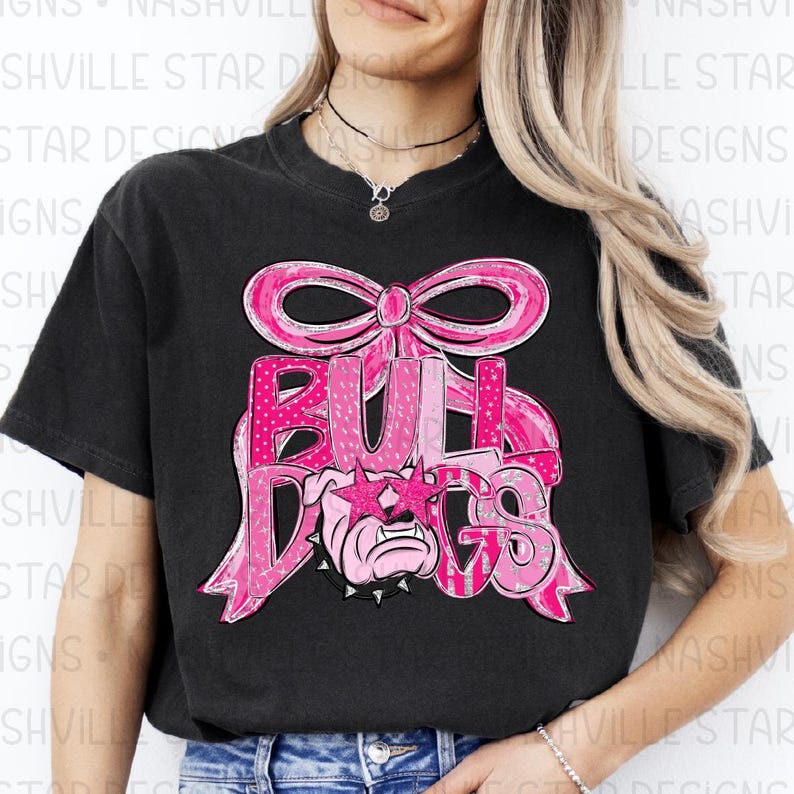May include: Black t-shirt featuring a pink graphic design. The design includes the word "BULLDOGS" in a stylized font, a bow, and a bulldog wearing star-shaped sunglasses. The graphic has a polka dot pattern.