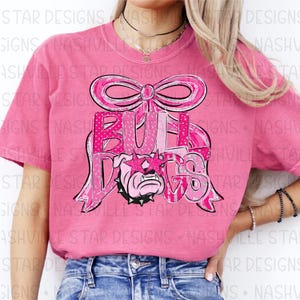May include: Pink t-shirt featuring a graphic of a bulldog wearing star-shaped sunglasses, the word "BULLDOGS" in pink letters, and a pink bow. The shirt is a solid pink color.