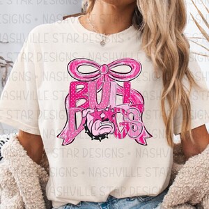 May include: Cream-colored t-shirt featuring a pink graphic design. The design includes the word "BULLDOGS" in a stylized font, a bulldog face, and a decorative bow. The shirt has a crew neck and short sleeves.