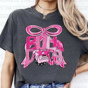 May include: Dark gray t-shirt featuring a pink bow and the word "BULLDOGS" in pink lettering. The design includes a bulldog wearing star-shaped sunglasses. The shirt is a casual style.