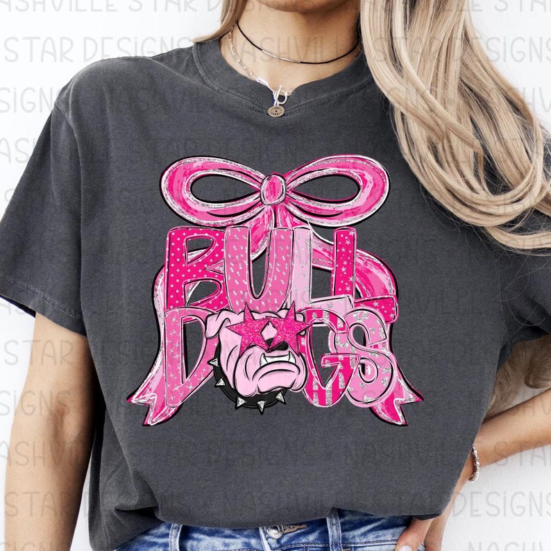 May include: Dark gray t-shirt featuring a pink bow and the word "BULLDOGS" in pink lettering. The design includes a bulldog wearing star-shaped sunglasses. The shirt is a casual style.
