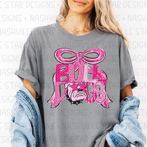 May include: Gray t-shirt featuring a pink bow and the word "BULLDOGS" in a pink, patterned font. The design includes a bulldog graphic wearing star-shaped sunglasses. The shirt has short sleeves.