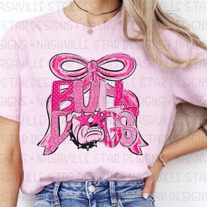May include: Pink t-shirt featuring a pink bow and the word "BULLDOGS" in a pink and silver design. The graphic includes a bulldog wearing star-shaped sunglasses. The shirt is a light pink color.
