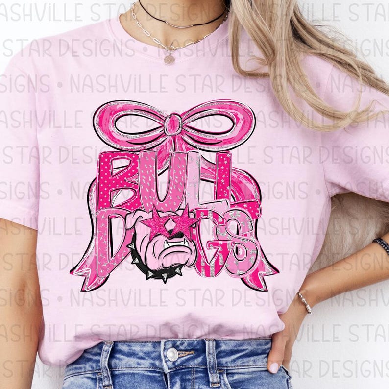 May include: Pink t-shirt featuring a pink bow and the word "BULLDOGS" in a pink and silver design. The graphic includes a bulldog wearing star-shaped sunglasses. The shirt is a light pink color.
