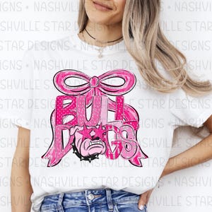 May include: White t-shirt featuring a pink graphic design. The design includes the word "BULLDOGS" in a pink, glittery font, a pink bow, and a bulldog wearing star-shaped sunglasses. The shirt is a casual style.