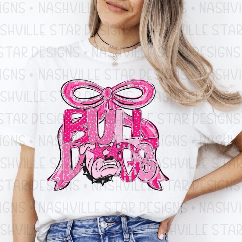 May include: White t-shirt featuring a pink graphic design. The design includes the word "BULLDOGS" in a pink, glittery font, a pink bow, and a bulldog wearing star-shaped sunglasses. The shirt is a casual style.