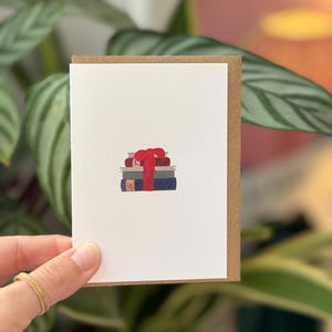 Puede incluir: Una tarjeta de felicitación con una pila de libros atados con una cinta roja. La tarjeta tiene una parte delantera blanca con un borde marrón. El fondo presenta follaje verde.
