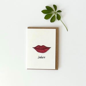 Può includere: Un biglietto d'auguri rettangolare con la parte anteriore bianca che presenta un'illustrazione disegnata a mano di labbra rosso scuro centrate sopra il testo nero J'adore. Il biglietto è abbinato a una busta in carta kraft marrone e una foglia verde.