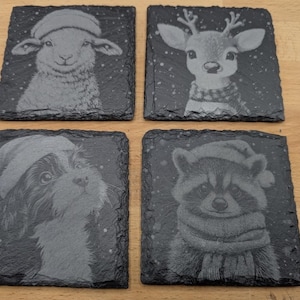 Puede incluir: Juego de cuatro posavasos cuadrados de pizarra con retratos de animales grabados. Incluye una oveja, un ciervo, un perro y un mapache, cada uno con un gorro y bufanda de Papá Noel. Los posavasos son de color gris oscuro.