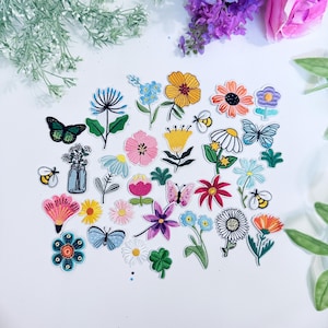May include: A collection of embroidered patches featuring various floral and insect designs. The patches include colorful flowers, butterflies, and bees in a variety of shapes and sizes. The patches are arranged on a white surface, with some greenery and flowers in the background.