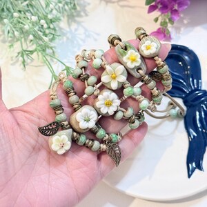 May include: Handmade bracelets featuring white flower accents, green beads, and bronze leaf charms. The bracelets are strung on beige cords and have a bohemian style. The floral elements add a touch of nature-inspired elegance to the jewelry.