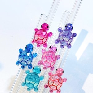 May include: Six clear glass straws adorned with colorful, glittery turtle charms. The turtles are in shades of pink, blue, and purple, with detailed shell patterns. The straws are arranged diagonally against a white background.