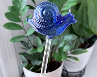 May include: A cobalt blue glass snail-shaped plant waterer. The snail has a textured shell and sits atop a clear glass stem. The plant waterer is designed to be inserted into the soil of a potted plant. The background includes green plants.