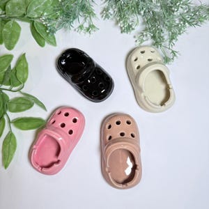 May include: Four ceramic ashtrays shaped like clogs in black, cream, pink, and tan. Each ashtray has a rounded shape with holes on top and a curved opening. The ashtrays are arranged on a white surface with green foliage.