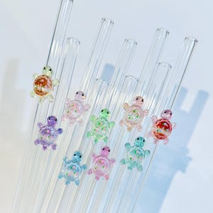 May include: Clear glass straws adorned with colorful, miniature turtle charms. The turtles are in various pastel colors, including pink, blue, green, and purple, with floral details. The straws are angled against a white background.