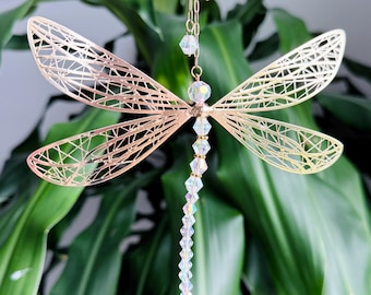 May include: A decorative dragonfly ornament with intricate, openwork wings in a gold tone. The dragonfly hangs from a delicate gold chain and features a beaded body of clear, faceted crystals. The background is a blurred view of green foliage.