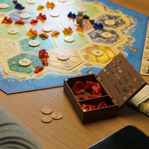 Op de afbeelding: Een bordspelopstelling met een zeshoekig spelbord, grondstofkaarten en spelstukken. Het bord heeft landtegels en een kustgrens. Rode, blauwe en gele spelstukken zijn zichtbaar. Een houten doos bevat rode spelstukken. Drie munten liggen op tafel.