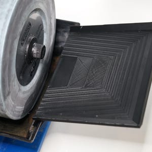 Könnte beinhalten: Nahaufnahme einer grauen Schleifscheibe, die an einer schwarzen und blauen Schleifmaschine befestigt ist. Die Schleifscheibe wird zum Schärfen von Werkzeugen und anderen Gegenständen verwendet. Die Maschine hat ein schwarzes Tablett mit einer strukturierten Oberfläche.