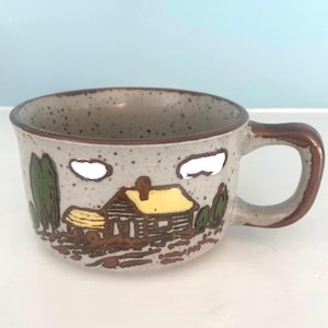 May include: A speckled ceramic mug with a brown handle and rim. The mug features a cabin scene with trees and clouds in shades of brown, yellow, and green. The mug is designed for hot beverages.