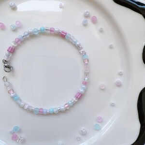 May include: A delicate bracelet made with small, colorful beads in shades of pink, blue, and white. The bracelet is made with a variety of bead shapes and sizes, creating a unique and eye-catching design.