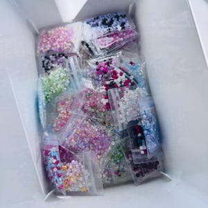 Puede incluir: Una colección de pequeñas cuentas de colores en bolsas de plástico transparente. Las cuentas son de varios colores, incluyendo rosa, morado, blanco, negro y verde. Las bolsas están dentro de una caja blanca, lo que sugiere que son para manualidades o bisutería.