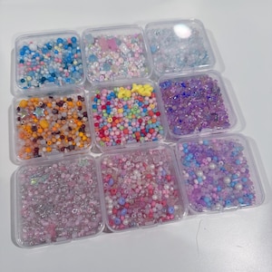 Op de afbeelding: Negen doorzichtige plastic containers gevuld met kleurrijke kralen in verschillende vormen en maten. De kralen zijn in tinten blauw, roze, paars, geel, oranje en wit. Sommige containers hebben ster- en vlindervormige kralen. Ideaal voor het maken van sieraden en knutselen.
