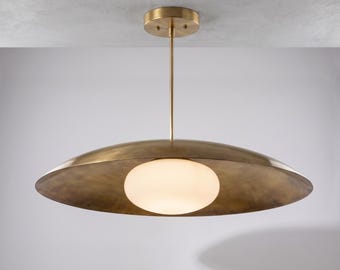 May include: A pendant light fixture featuring a wide, shallow, brushed brass metal shade with a smooth, matte finish. A central, spherical, frosted white glass globe is suspended from a thin brass rod attached to a circular ceiling canopy.