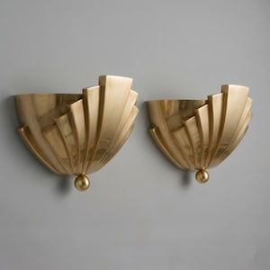 May include: A pair of gold-toned wall sconces, each featuring a fan-shaped design. The sconces have a rounded base with a small sphere and a series of stepped, vertical elements. They are mounted on a grey wall, creating a striking contrast.