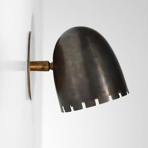 May include: A vintage wall sconce with a dark, metallic finish. The lamp has a rounded shade with a slotted bottom edge and a circular mounting plate. The fixture is mounted on a white wall.