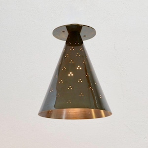 Puede incluir: Una lámpara de techo de latón con una pantalla en forma de cono. La pantalla tiene un fondo de color cobre y está perforada con pequeños agujeros. La luz está encendida y los agujeros están iluminados.