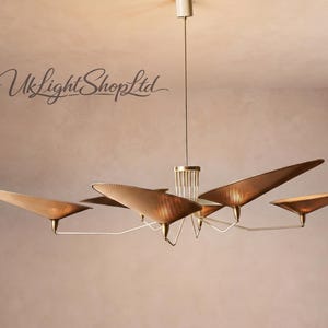 May include: A multi-arm chandelier featuring six triangular, metallic gold-toned shades extending from a central vertical column. The fixture is suspended by a thin rod and includes the text UkLightShopLtd in a cursive script in the upper left corner.
