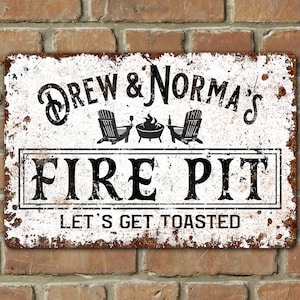 May include: White metal sign with black text that reads "Drew & Norma's Fire Pit Let's Get Toasted". The sign features a silhouette of two Adirondack chairs and a fire pit with flames.